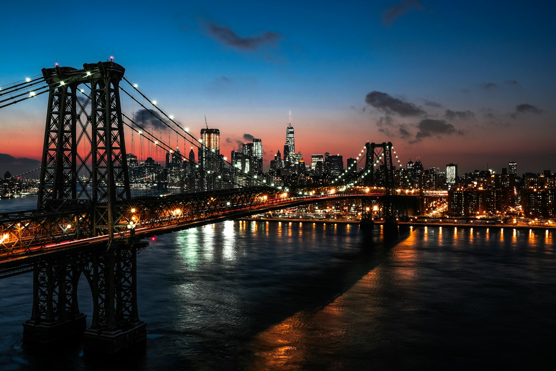 Manhattan skyline lit up at night behind the Williamsburg Bridge, with city lights reflecting on the East River